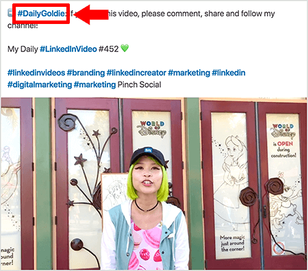 Dette er et skjermbilde som illustrerer hvordan Goldie Chan bruker hashtags i teksten til sine LinkedIn-videoinnlegg. Røde forklaringer peker på #DailyGoldie-hashtaggen i teksten, som er unik for hennes videoinnlegg og hjelper henne med å spore aksjer. Innlegget inneholder også andre relevante hashtags som hjelper folk med å finne videoen hennes, inkludert #LinkedInVideo. I videobildet står Goldie foran noen dører på en World of Disney-skjerm. Hun er en asiatisk kvinne med grønt hår. Hun har på seg en svart LinkedIn-hette, et svart choker-halskjede, en rosa skjorte med makaron-trykk og en blå og hvit jakke.