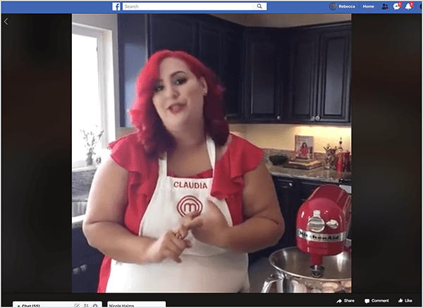 Dette er et skjermbilde av en Facebook Live-video med kokk Claudia Sandoval, som hun var vertskap for i 2016 som en del av en krysskampanje med T-Mobile Tirsdager-appen. I videoen står Claudia på et kjøkken med sorte skap og benkeplater i granitt. Et vindu over vasken slipper naturlig lys inn i rommet. Claudia står ved siden av en rød Kitchen Aid-mikser. Hun er en latinsk kvinne som har knallrødt hår som faller rett under skuldrene hennes. Hun har på seg sminke, en lys rød topp og et hvitt forkle med Claudia og MasterChef-logoen brodert i rød tråd. Mens hun snakker, gestikulerer hun med hendene. I 2016 samarbeidet Stephanie Liu med Claudia for å produsere denne livevideoen.